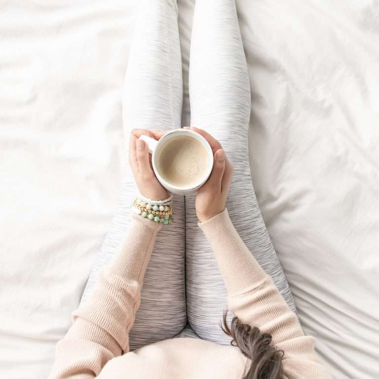 Shot from above. A woman is sat on a white bed. She is wearing grey leggings, a pink jumper and she is holding a coffee cup