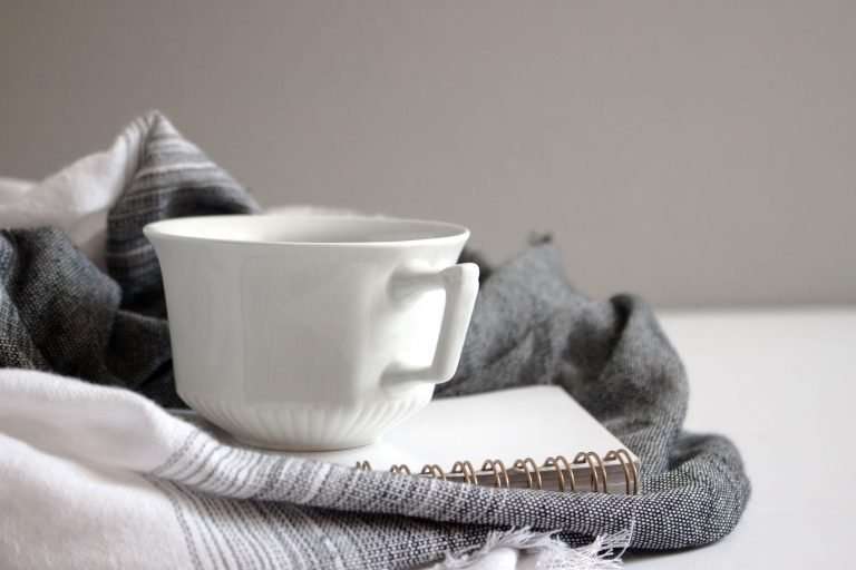 A white teacup is resting on a white notebook and grey and white blanket