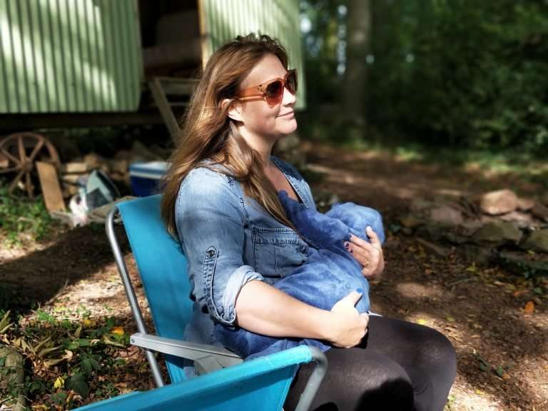 A woman is sat outside on a deck chair. She is wearing sunglasses and a denim shirt. She is breastfeeding her baby