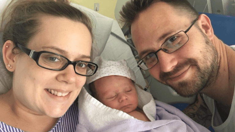 Lisa and Darren smiling at the camera. Little J is a newborn and has his eyes closed. They are in a hospital bed and Lisa is wearing a hospital gown