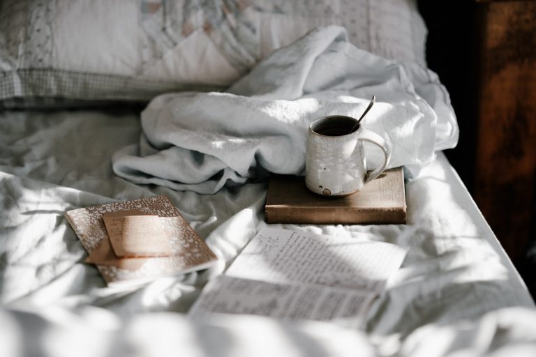 An unmade bed with white and grey bed sheets. Books, a note pad and a coffee cup are resting on the bed