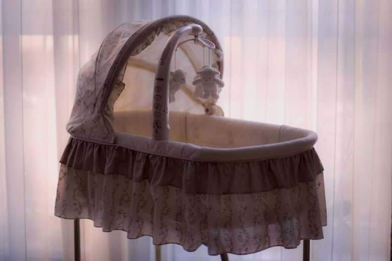 A Moses basket with a frilly covering. The hood is up and it is in front of a net curtain. There is a lamb mobile handing above it