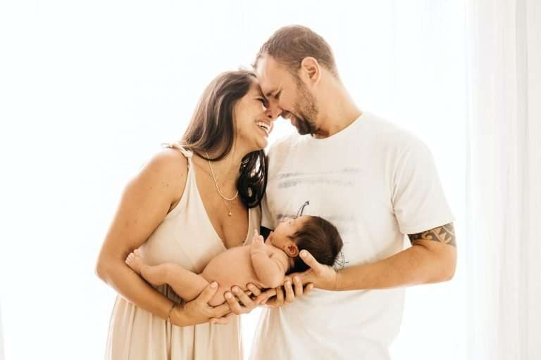 A man and a woman are smiling at each other and resting their heads together. They are holding a newborn baby