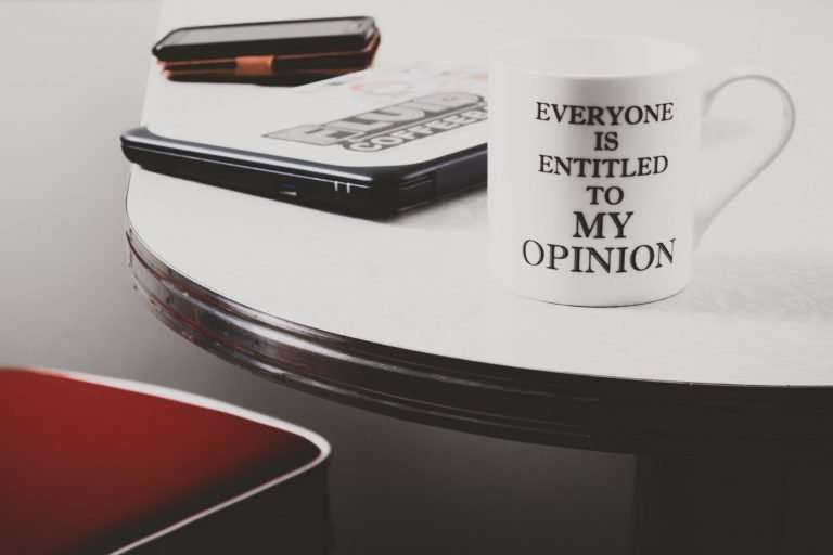 A white table has a laptop, phone and mug on it. On the mug is written 'everyone is entitled to my opinion. There is a red chair next to the table.