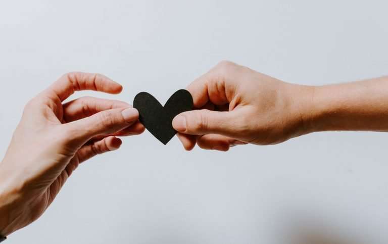 A plain white/grey background. There are two hands and it looks as though one is passing the other a black paper heart