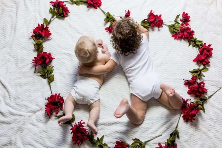 A baby and toddler are laying on their fronts on a white bed. The toddler has their arm around the the baby. The baby is in a white nappy and the toddler is wearing a white baby grow. Around them is lots of red flowers in the shape of a heart