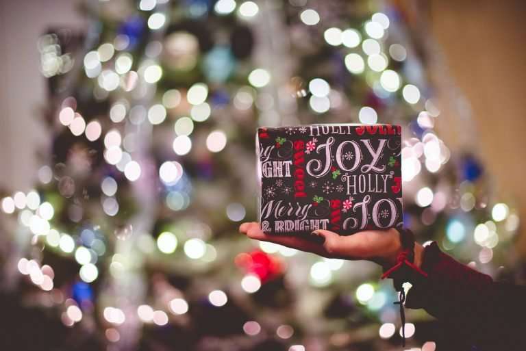 A hand is holding up a single present in their hand. It is wrapped in black paper and has red and white writing on it. In the background is an out of focus Christmas tree