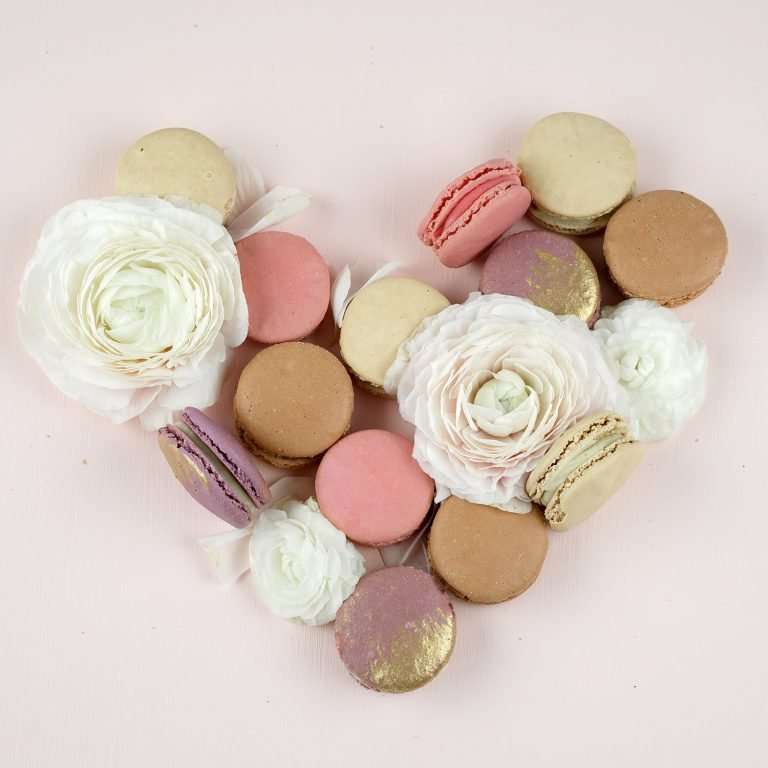 A pale pink background. White flowers and pink, brown, purple and white macarons are making a heart