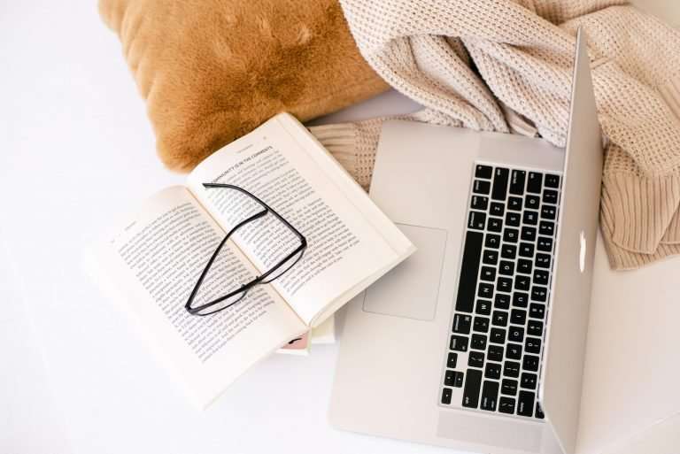 There is a laptop on a white surface. Next to it is a beige jumper and brown pillow. There is a book with a pair of glasses resting on them
