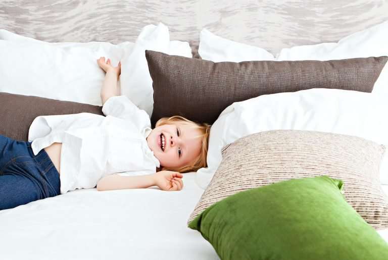 A toddler in jeans and a white shirt is laying on a large white bed. There are brown, beige and green cushions and the toddler is smiling