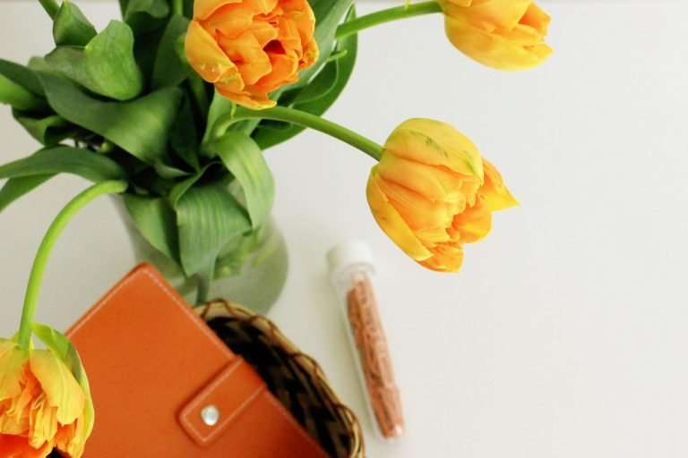 Shot of droopy yellow tulips from above in a vase. There is a brown dairy in a wicker basket and a test tube of bronze coloured paperclips
