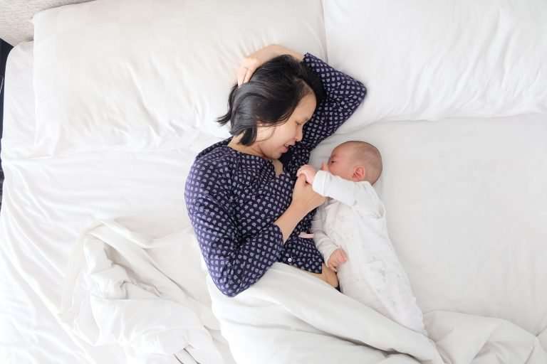 A mother and baby are laying in a white bed. The mother is wearing blue pyjamas. The baby is facing her and she is holding the baby's hand