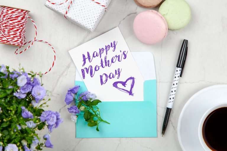Happy Mother's Day postcard in purple writing on a blue envelope on a white marble table. There are purple flowers on the table, a cup of coffee and macaroons