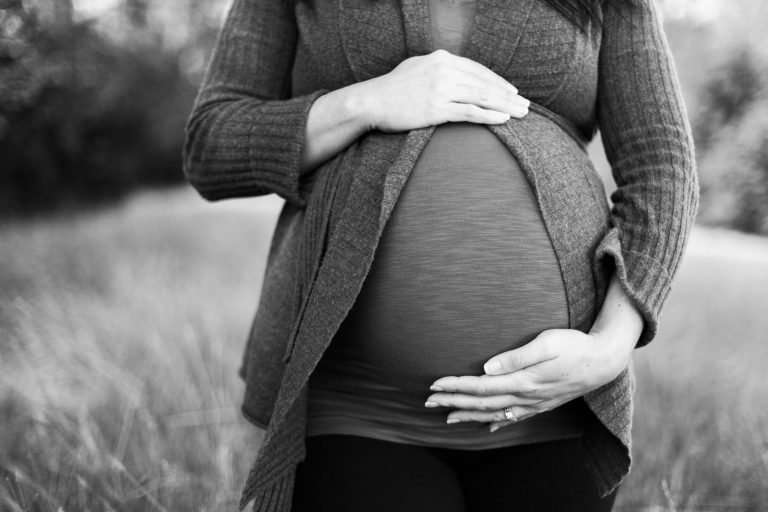 A black and white close up picture of a pregnant belly. The woman is cradling her bump with one hand at the top of the bump and one hand on the bottom