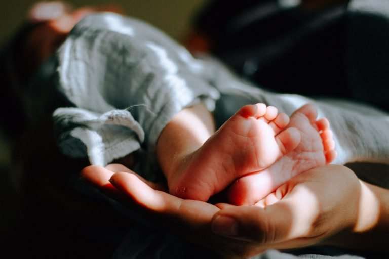 Close up of a hand. Resting in the hand are newborn baby feet. There is a blue blanket over the baby's legs