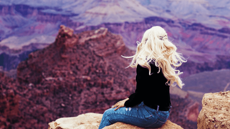The back of a woman with long blond hair, wearing jeans and a black jumper is sitting on top of a mountain looking out to other mountains