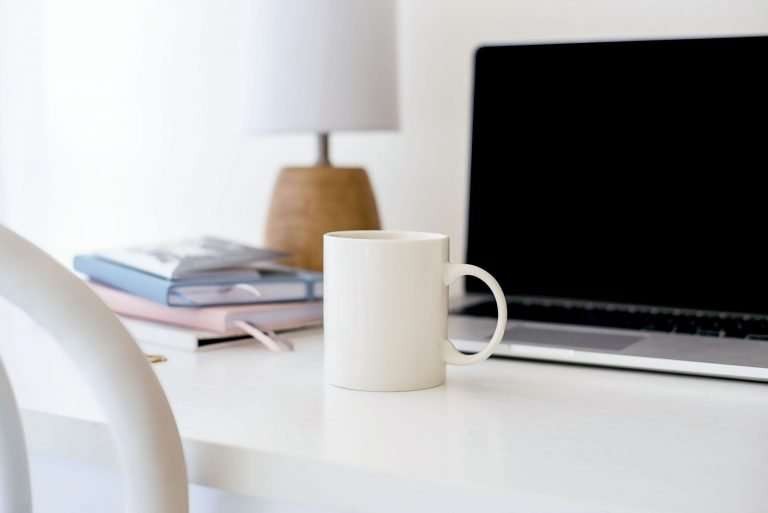 A white desk has a laptop on it. There is a white coffee cup, a blue notebook, a pink notebook and a white lampshade with a wooden base