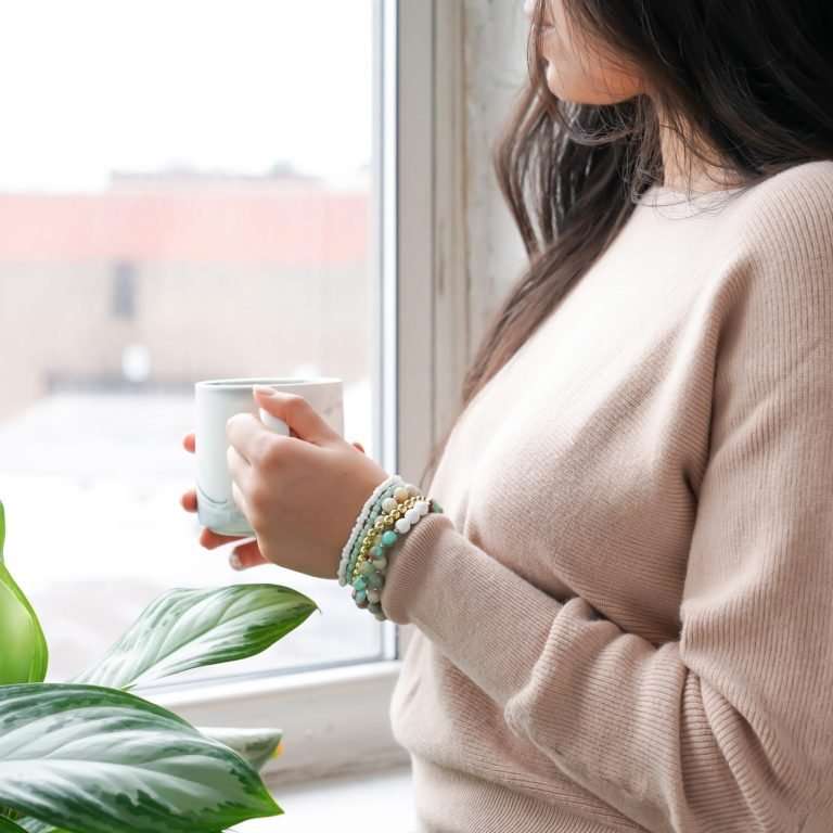 A woman in a pink jumper is holding a coffee mug and looking out the window. She has gold and green bracelets and there is a green plant next to her