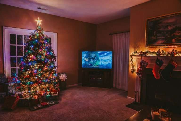 A cosy living room with a fireplace, stockings and a Christmas tree. The Grinch is playing on the TV. It is only lit by fairy lights