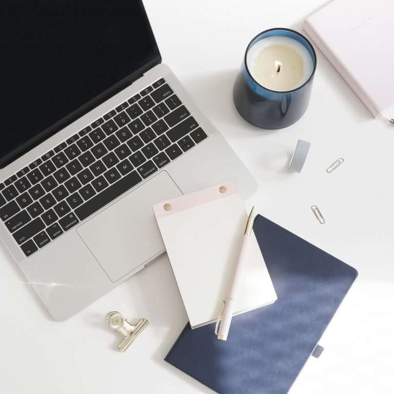 White desk with a laptop, paperclips and a note pad. There is a blue folder on the desk and a white candle in a glass blue holder