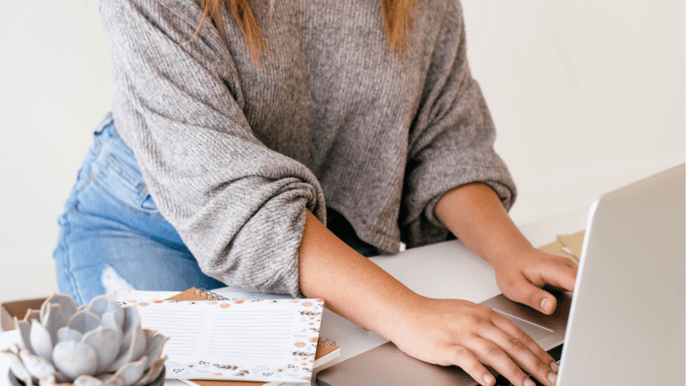 A woman in a grey jumper and jeans is sitting at a desk. She is working on a laptop and has a plant and paper on the desk