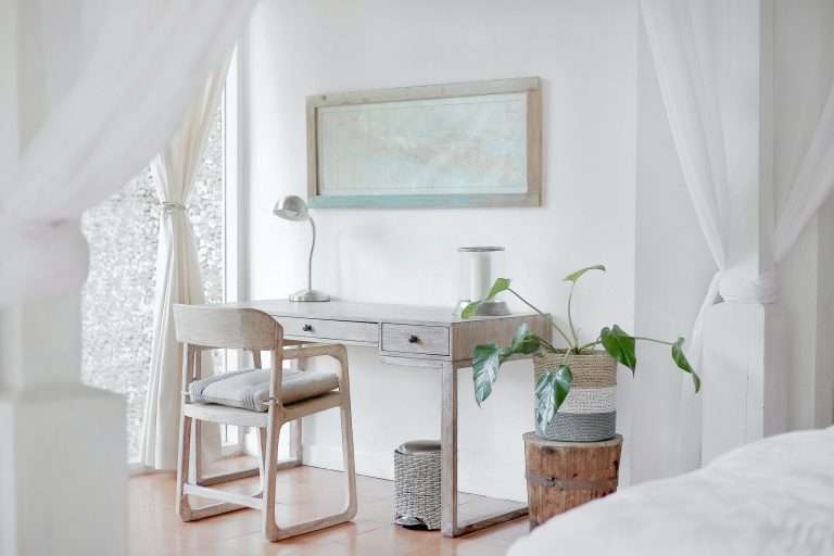 A white bedroom/office with white curtains hung up. There is a pale wooden desk with a lamp on it. A plant pot with a green plant is next to the desk