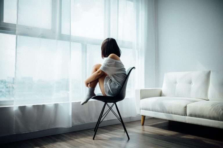A woman is sat on a hard black chair in front of a large window with a white net curtain. There is a soft white sofa next to her