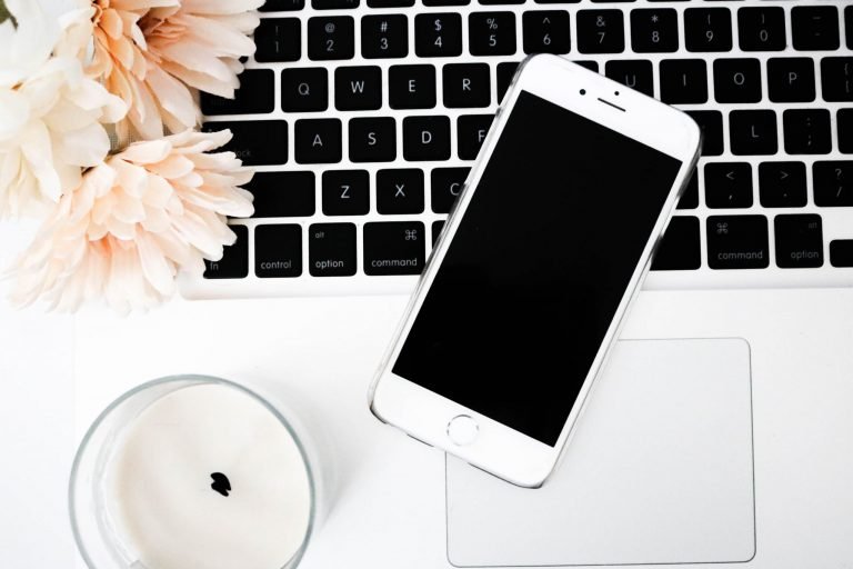 An iPhone is resting on a computer keyboard. there is a candle and the edge of some pink flowers