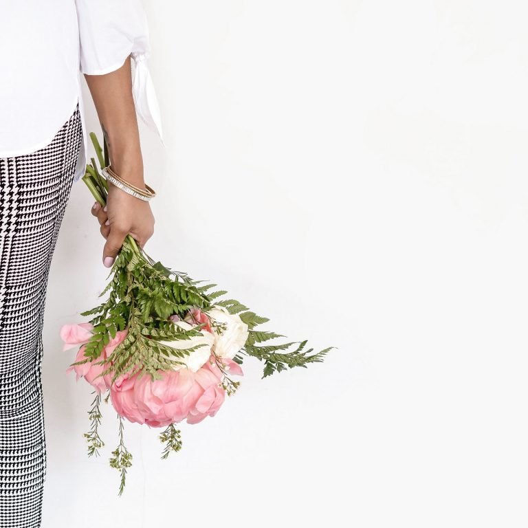 Can see left side only, waist down of a woman. She is wearing black and white checkered trousers and a white blouse. The sleeve goes to just below her elbow. She has a sparkly bracelet on and is holding a bunch of pink and white flowers with green leaves. The flowers are pointing down