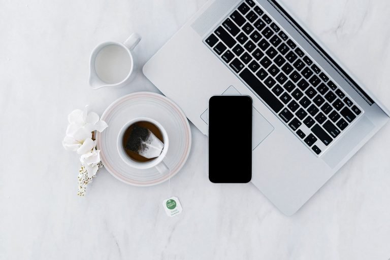 On a white marble table is a laptop, a white cup with black tea and tea bag with string on it, a white jug of milk, a mobile phone resting on the laptop keyboard, and a white flower