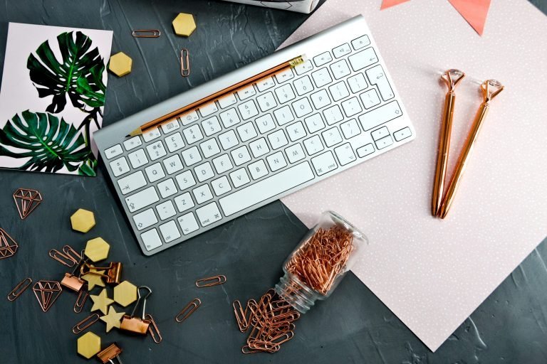 On a black desk is a computer keyboard. There is pink paper with white dots all over it, and two rose gold pens on top. There is a small jar that has been knocked over, and rose gold paperclips are spilling out. There is also a pink piece of card with large green leaves on it