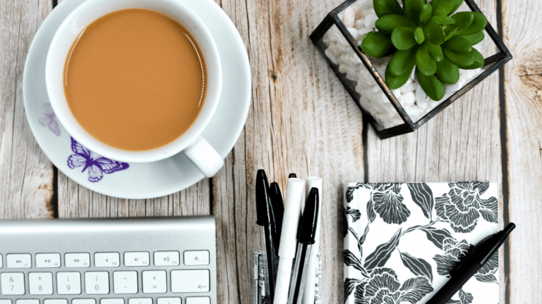 On a wooden table is a white cup and saucer with a purple butterfly on the saucer. The cup has a strong cup of tea in it. Also on the table is the edge of a computer keyboard, black and white pens, a black and white notebook and a Succulent plant in a pot with white pebbles