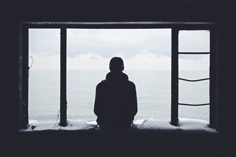 A man is sitting on a snowy ledge. His back is to the camera and he is looking out to sea