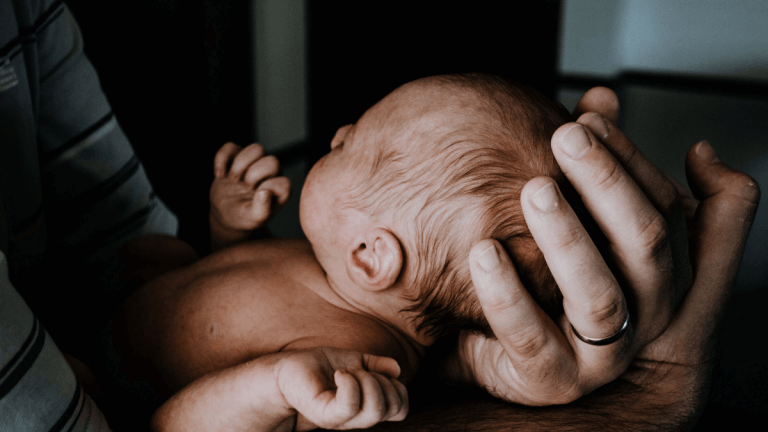 A newborn baby is being cradled by its dad. The dad is wearing a wedding ring