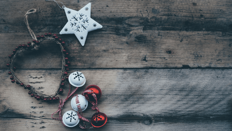 A heart-shaped Christmas decoration with white and bells attached to it sat on an old wooden table. There is also a star-shaped decoration on the table with snowflakes cut out of it