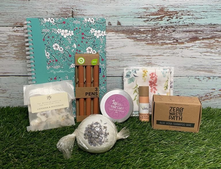Mental Health Awareness Week Giveaway goodies. Resting on grass and a blue wooden fence background are; Sunset Bath Bomb Aloe Vera Shampoo Bar Clearing Pink Clay Mask - 100% Natural Pink Clay Mask Powder Eco-Friendly Vegan Lip Balm In Sweet Orange Wax Melt in Lavender & Petitgrain Reusable Cotton Pads Journal and pens