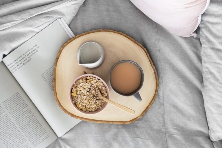 On a grey bed is an open book. There is a round wooden tray on the bed with a pink bowl full of oats with a wooden spoon, a jug of milk and a grey mug with tea in it