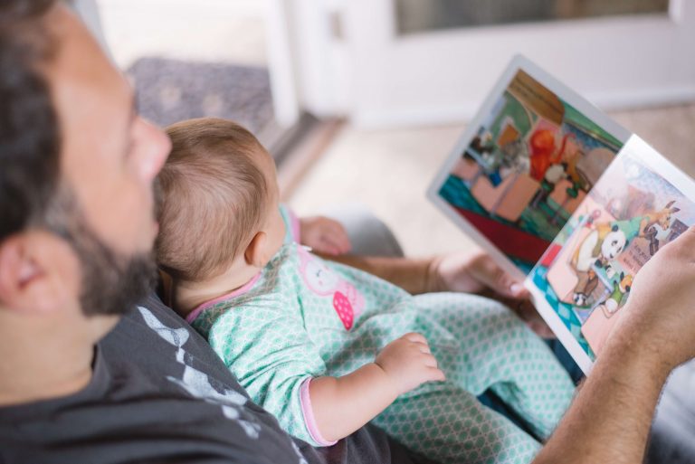A father in a grey top has a baby resting on his lap. The baby is in a green and white baby grow. The dad is holding a book and reading to his baby