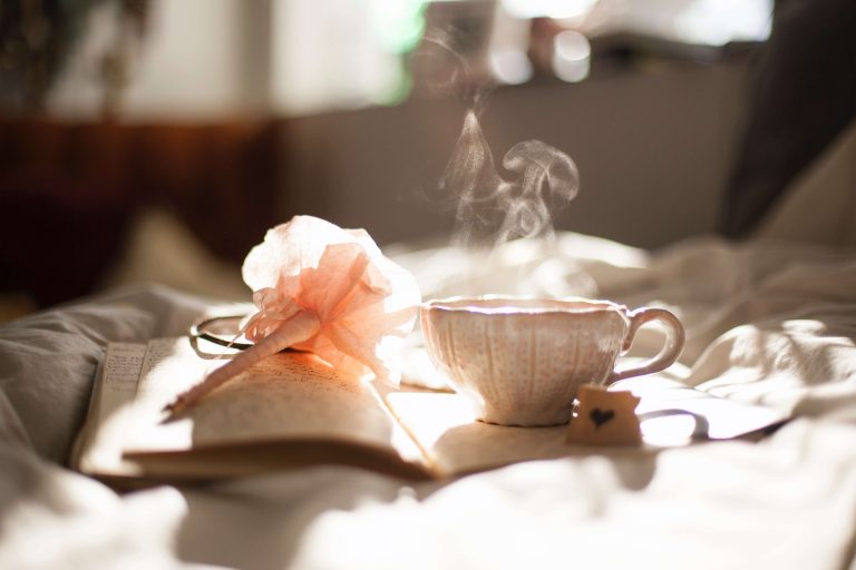 A small teacup is resting on a book. There is steam coming out of the cup. There is a pen with a flower topper also on the book