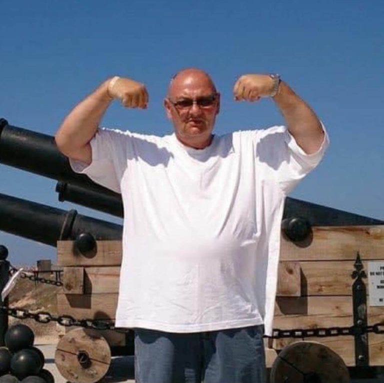 Ian is wearing blue shorts and a white t-shirt. He is tanned and the sky is blue. He is stood in front of canons and is pulling a pose like 'suns out guns out'