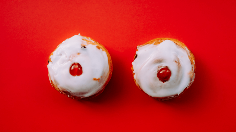 On a red table are two Belgian buns with red cherries on top. The image is designed to look like two breasts