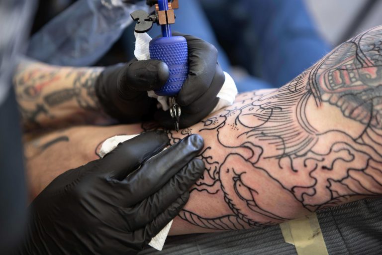 A person wearing black gloves is holding a blue tattoo machine over a persons arm. The person has the black outline of the tattoo