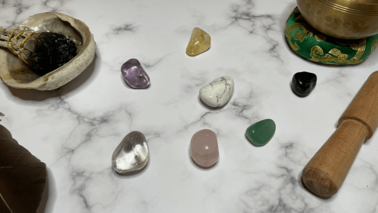 Seven different healing crystals are resting on a marble background. Around the crystals are sage in a shell, a feather and a singling bowl resting on a green and gold cushion