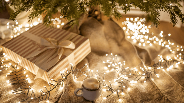 On a beige coloured rug, underneath a Christmas tree, is a Christmas present in stripey pink and white paper. There is a white bow wrapped around the present. There is a mug of frothy hot chocolate next to it and a string of fairy lights on the floor around the tree