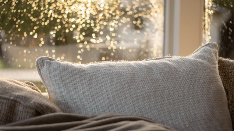 A beige/off white pillow is on a brown sofa. There is a window behind it. Sunlight is shining on the rain drops on the window to make it look like drops of gold