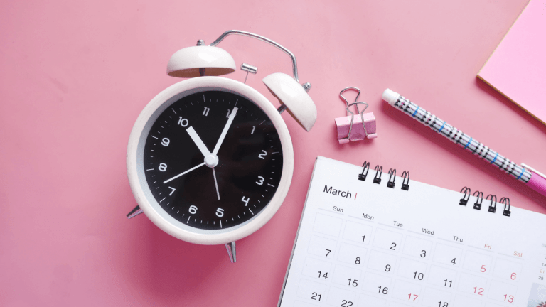 On a very pink table is a soft pink alarm clock showing 10 o'clock. There is a calendar on the table showing March. There is a tartan pink and blue pen, a. pink bulldog clip and some pink paper.