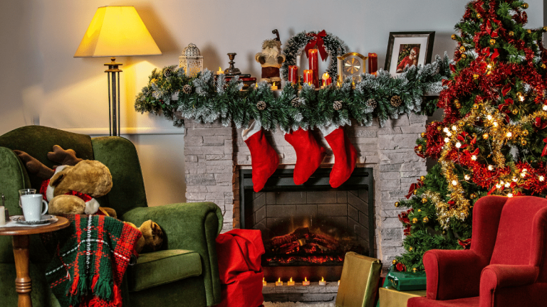 A living room is heavily decorated with Christmas decorations. There is a Christmas tree with red and gold tinsel and lights, there are 3x stockings hanging over the fire place and lots of candles on the fireplace, along with a garland. There is a soft toy reindeer sat in an arm chair