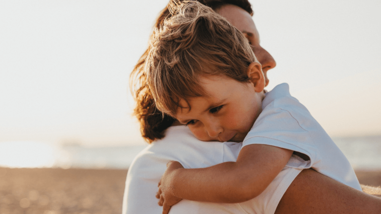 A little boy in a white shirt is hugging his mother. She is also in a white shirt and they are on the beach
