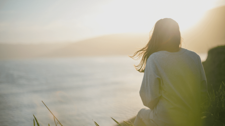 A woman with long hair blowing in the wind is sitting on a cliff overlooking the sea. Her back is to the camera, and she is wearing a white cardigan