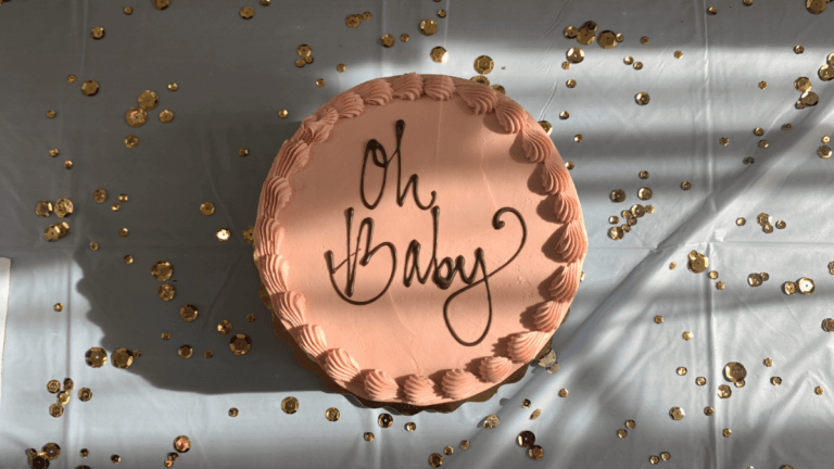 A pastel orange cake has Oh Baby written on it in chocolate. It is sat on a grey table cloth and there is gold confetti over the table